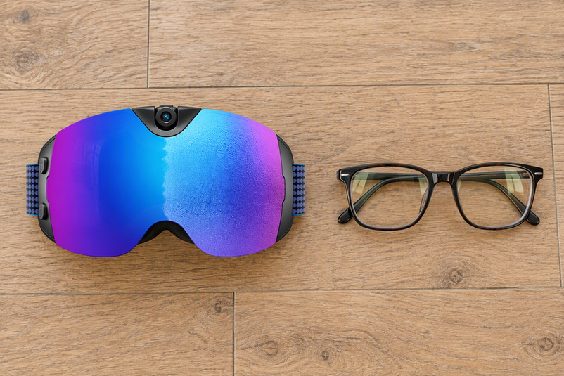 Skyshot camera ski goggles with a pair of prescription glasses set beside them, not inside the goggles